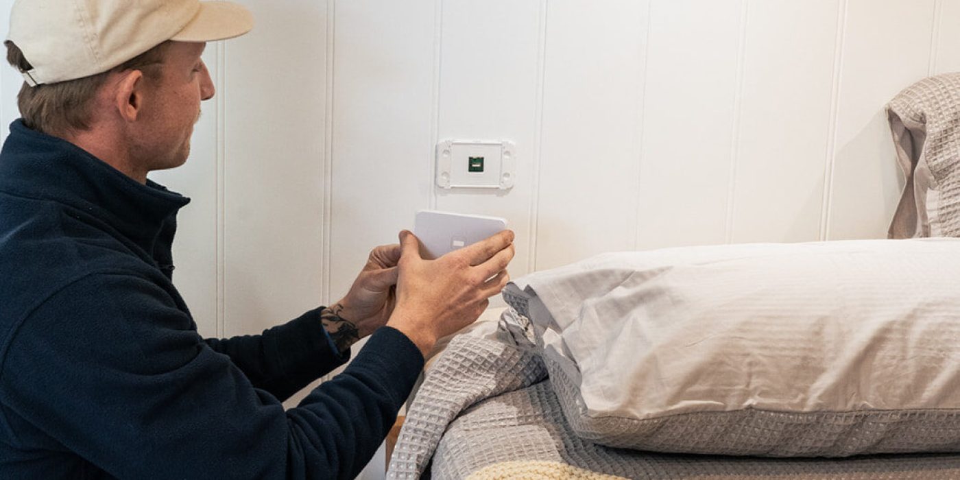 Electrician installing air conditioning system near bed in Perth home setting.